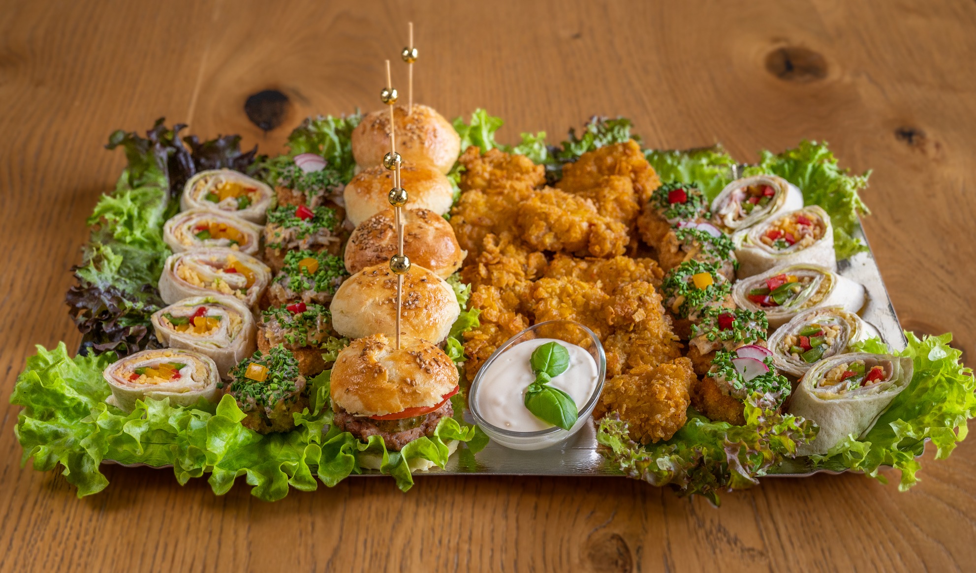 Elegancki srebrny półmisek z różnorodnymi przekąskami: mini burgery z dekoracyjnymi wykałaczkami, tortille w plastrach z kolorowym nadzieniem, nuggetsy i przystawki udekorowane ziołami, podane...