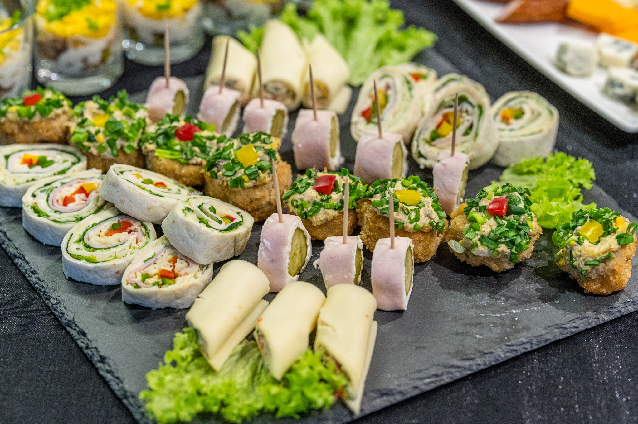 Elegancko ułożone na czarnym kamiennym talerzu przekąski cateringowe: roladki z tortilli z szynką i warzywami, koreczki z szynką i ogórkiem, grzanki z pastą jajeczną i szczypiorkiem, sałatki...