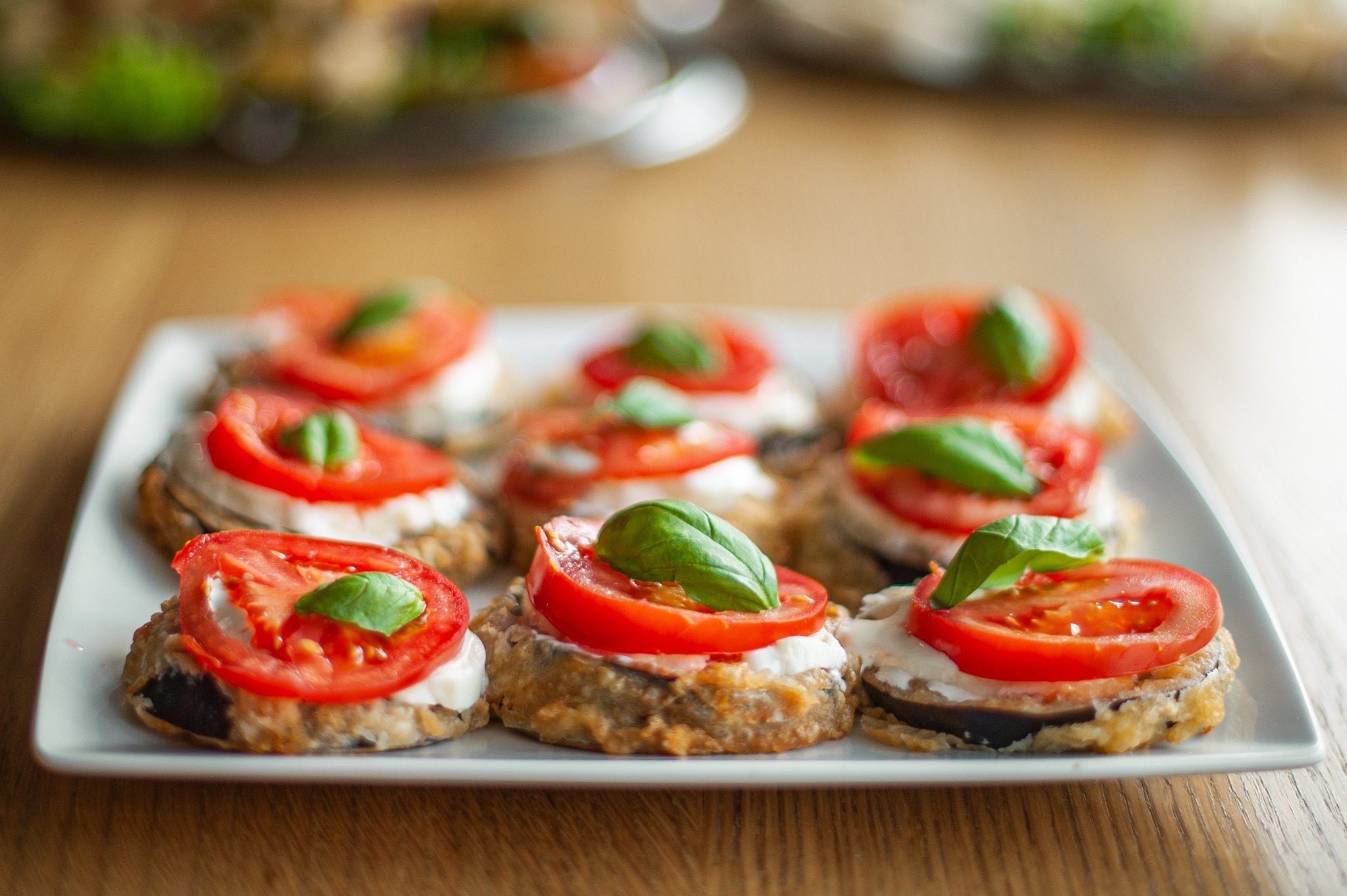 Apetizing koreczki z bakłażana, pomidora i bazylii na białym talerzu, na drewnianym stole, w tle inne przekąski.