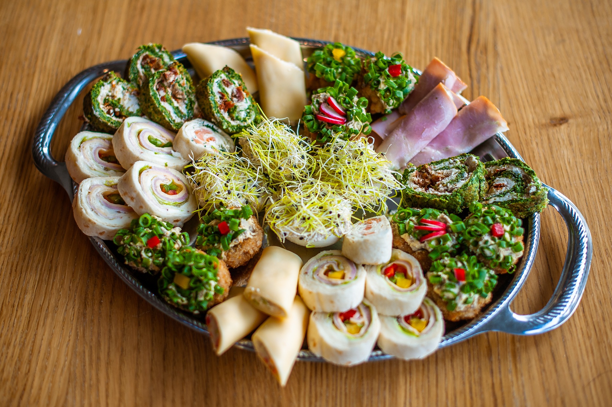 Dekoracyjna srebrna taca z różnorodnymi przekąskami: roladki z szynką i warzywami, koreczki z pastą i szczypiorkiem, roladki ze szpinakiem i suszonymi pomidorami, plastry sera i szynki, całość...