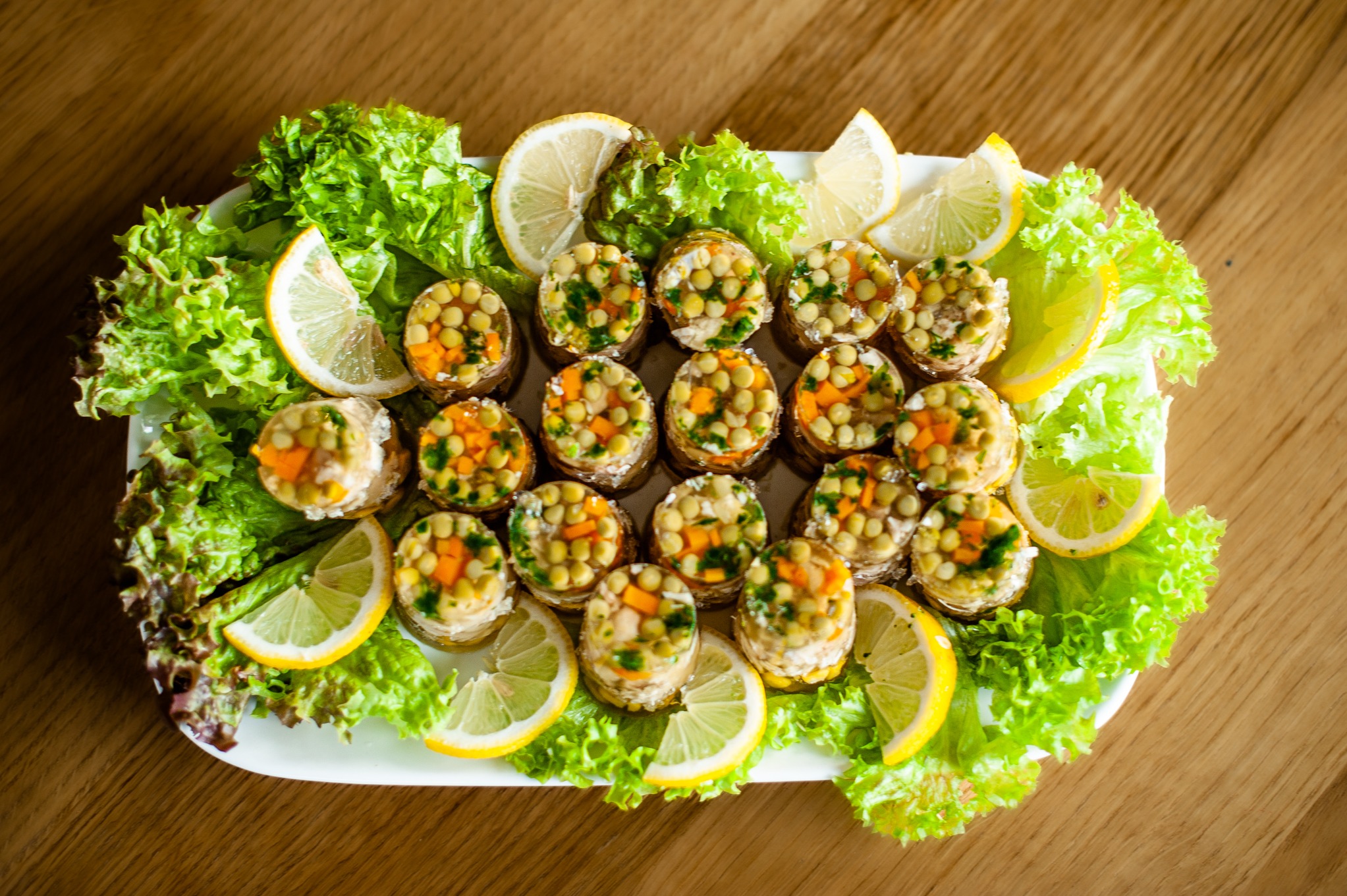 Dekoracyjnie ułożone galaretki z groszkiem i marchewką na białym talerzu, otoczone świeżą sałatą i plasterkami cytryny, na drewnianym stole.