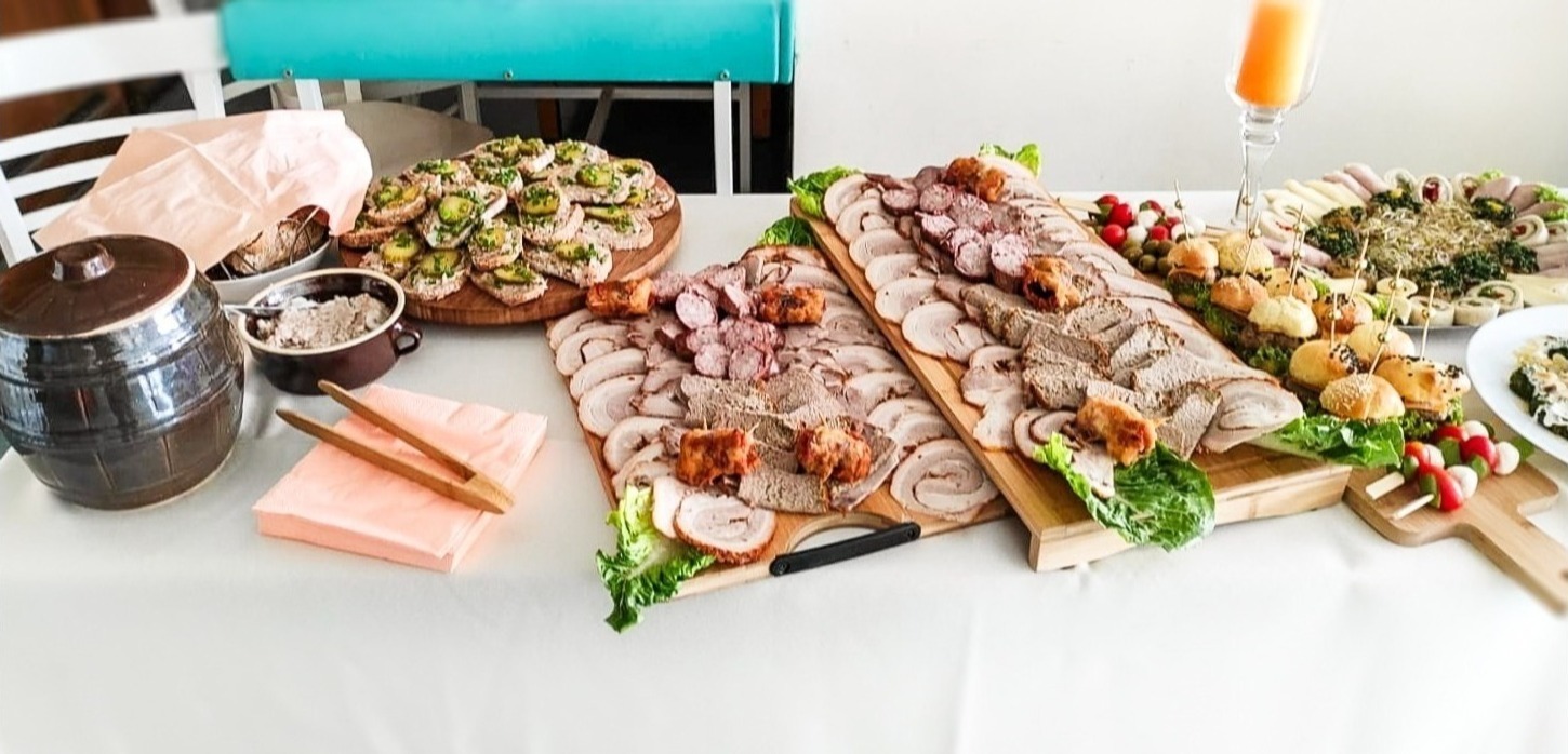 Bogato zastawiony stół z deskami wędlin, kanapeczkami z pastą i ogórkiem, mini burgerami, szaszłykami i roladkami, uzupełniony o gliniany garnek i słoik z sokiem.