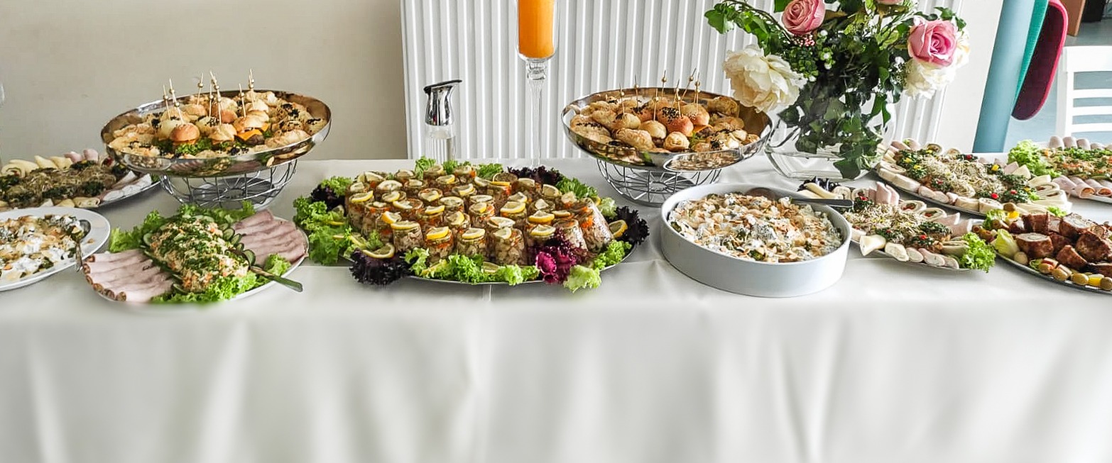 Stół bankietowy z różnorodnymi potrawami: mini burgery na srebrnej paterze, galaretki z cytryną, sałatki, roladki z szynki i mięsa, dekoracje z kwiatów i soku pomarańczowego.