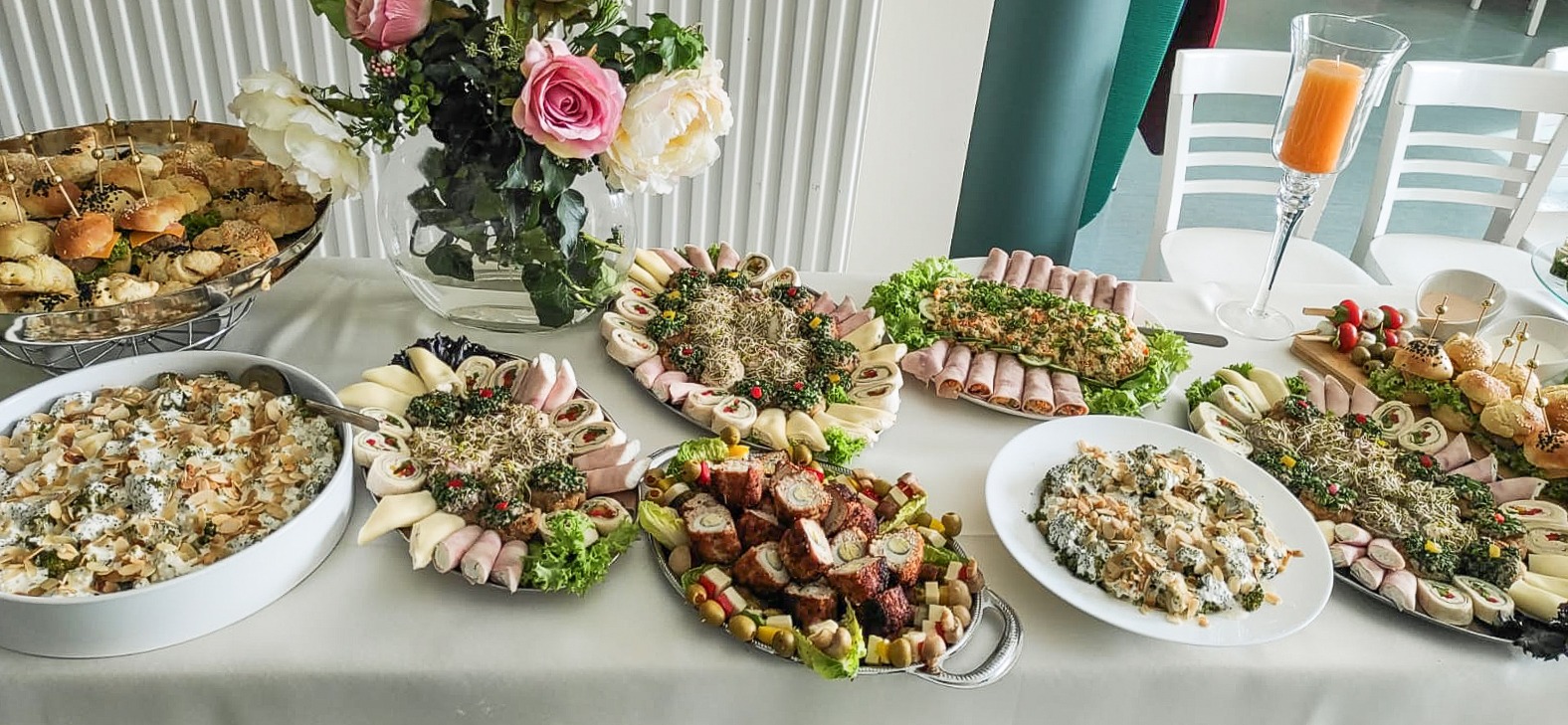 Bogato zastawiony stół z różnorodnymi przekąskami: mini burgery na szpilkach, sałatki warzywne z migdałami, roladki z szynki i sera, koreczki z pomidorkami i oliwkami, dekoracje kwiatowe w szklanym...