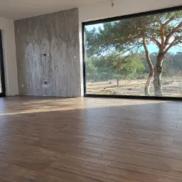 Ułożona podłoga z paneli imitujących drewno w jasnym, przestronnym pokoju z dużymi oknami wychodzącymi na las. Na ścianie widoczna szara faktura betonu i puszki elektryczne.