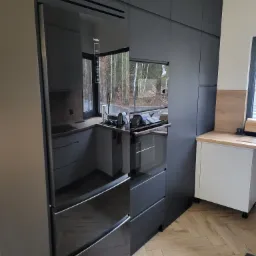 Nowoczesna kuchnia z zabudową w kolorze antracytowym, zintegrowana lodówka Haier z czarnym frontem, parkiet ułożony w jodełkę, widok na okno i zieleń za nim.