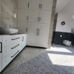 Biała zabudowa meblowa w łazience z szafkami i szufladami z czarnymi uchwytami, marmurowe płytki na ścianie, widoczna toaleta w tle, zabezpieczona podłoga podczas remontu.