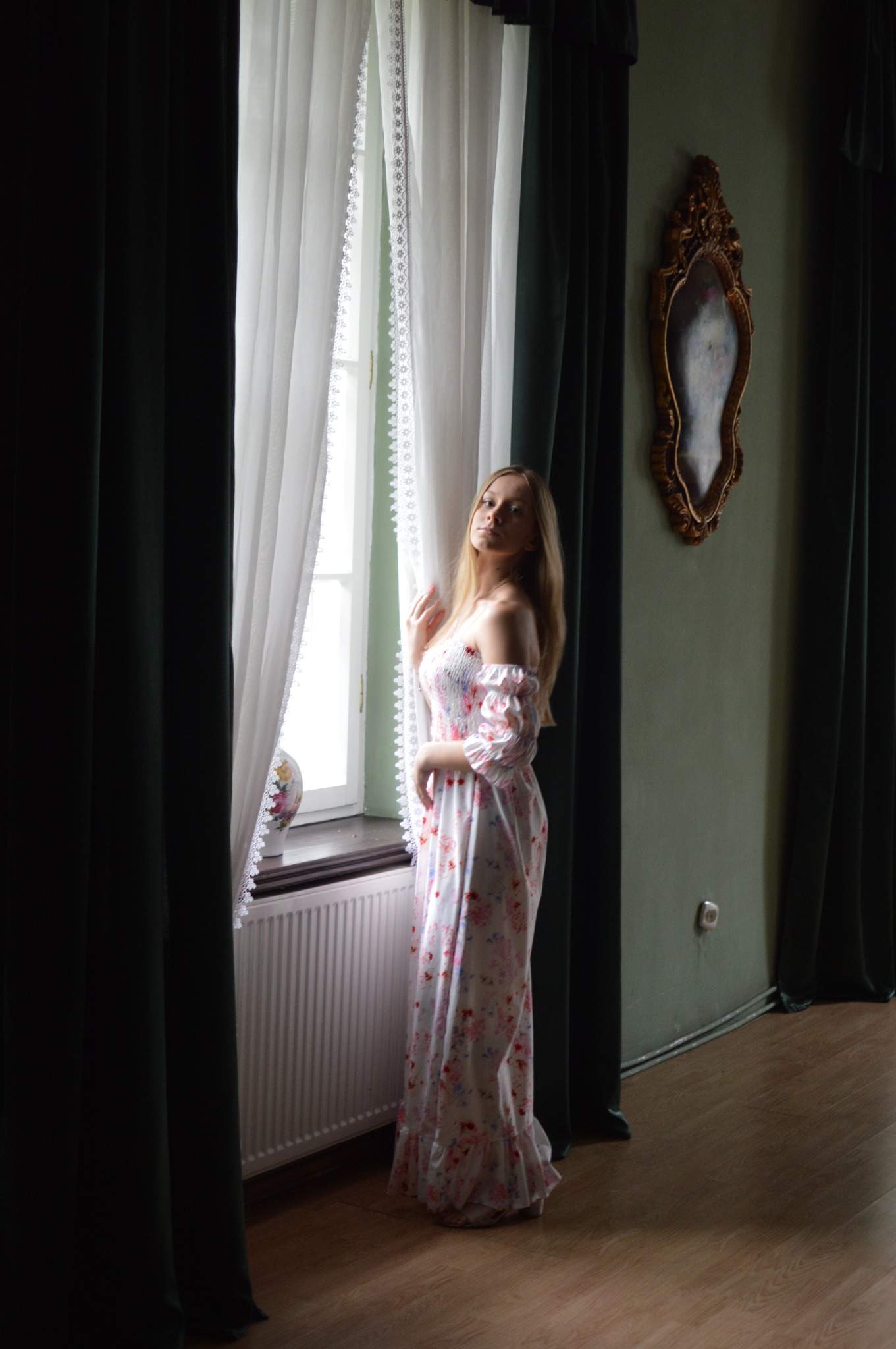 Kobieta w kwiecistej sukience stoi przy oknie z koronkowymi firankami w pokoju z zielonymi ścianami i złoconym lustrem.