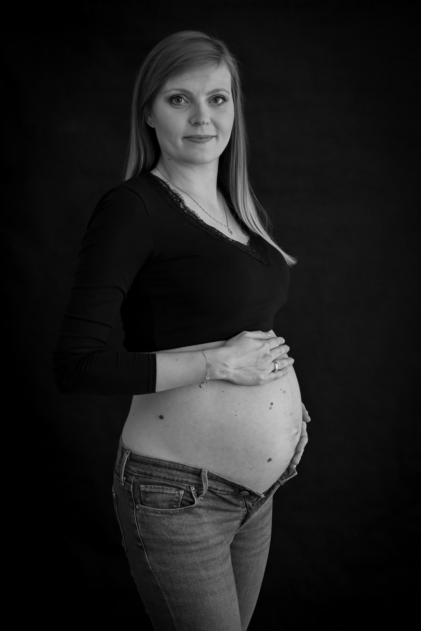 Czarno-białe portretowe zdjęcie kobiety w ciąży, ubranej w czarną bluzkę i dżinsy z rozpiętym guzikiem, dłonie spoczywają na brzuchu, lekki uśmiech, ciemne tło.