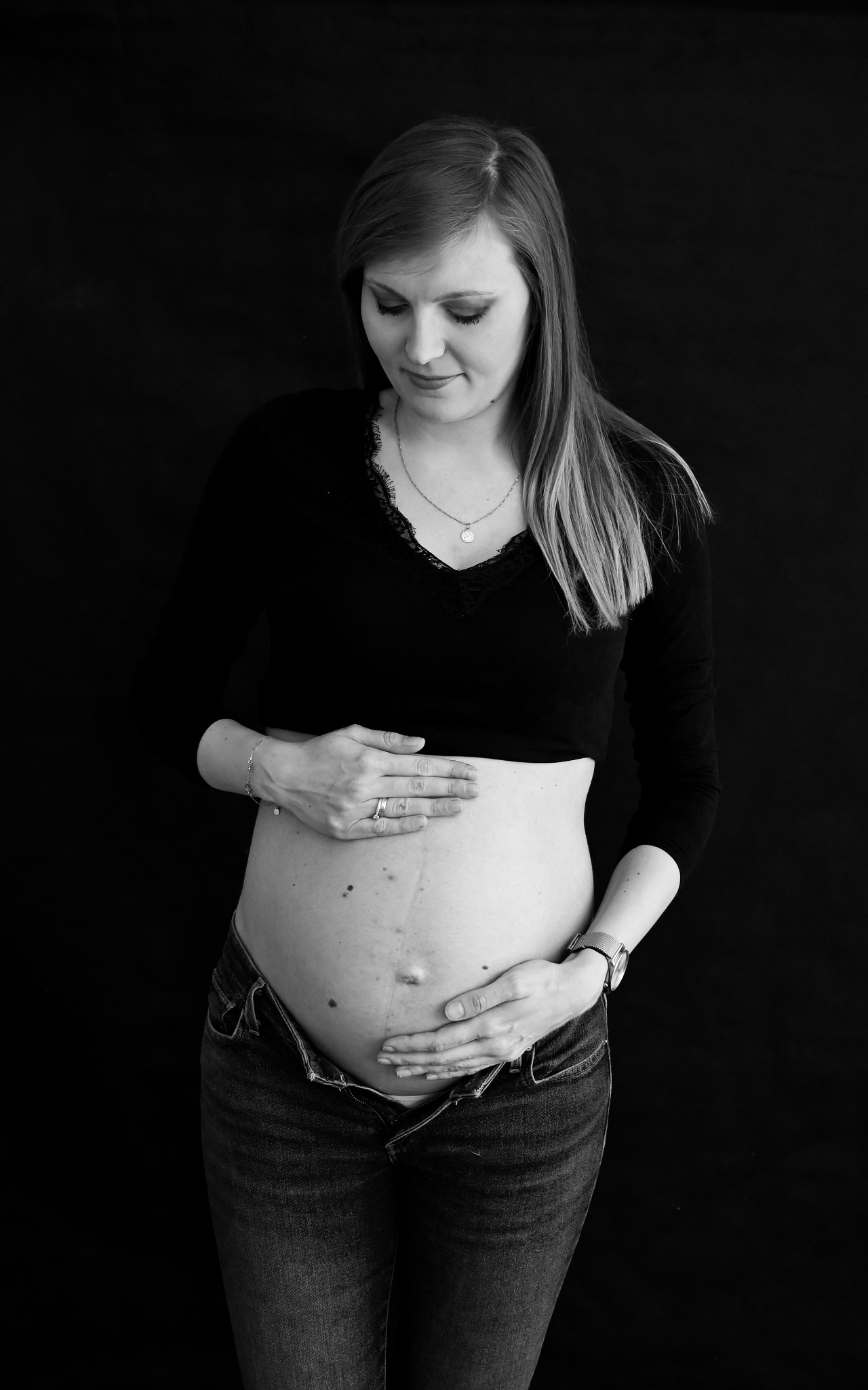 Czarno-białe zdjęcie kobiety w ciąży, z rękami na brzuchu, ubrana w czarną bluzkę z koronką i rozpięte dżinsy, na ciemnym tle.