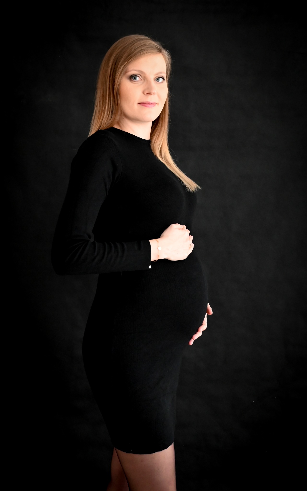 Portret kobiety w czarnej sukience, trzymającej ręce na brzuchu, na ciemnym tle. Widoczny subtelny bransoletka na nadgarstku.