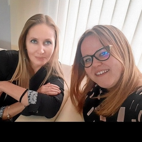 Dwie uśmiechnięte kobiety siedzące na jasnej kanapie, jedna z blond włosami i czarnym żakiecie, druga w okularach i bluzce w białe wzory na czarnym tle, za nimi białe żaluzje.