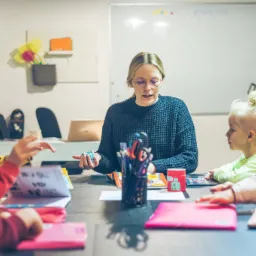 Nauczycielka w okularach prowadzi lekcję z dziećmi przy stole, na tablicy napis 'ENGLISH', widoczne materiały edukacyjne i przybory.