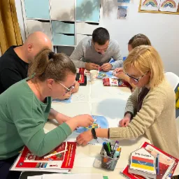 Grupa osób przy stole, pracująca nad mapą z flagami państw Ameryki Południowej; widoczne podręczniki do nauki języka hiszpańskiego, kubek i przybory do pisania.