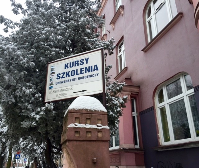Zimowy widok na tablicę informacyjną z napisem 'Kursy Szkolenia Uniwersytet Robotniczy', umieszczoną przed budynkiem uczelni. Na tablicy adres 'ul. Sienkiewicza 9, II piętro'.