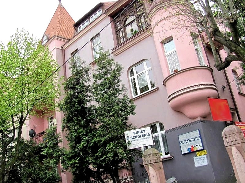 Elewacja budynku w pastelowych kolorach z szyldami informującymi o kursach i szkoleniach, widoczne drzewa i elementy architektoniczne.