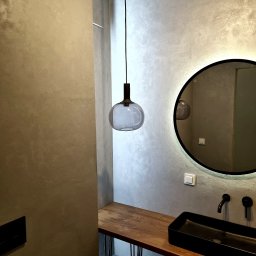 ALFA-BUD KM SPÓŁKA Z OGRANICZONĄ ODPOWIEDZIALNOŚCIĄ - Szara ściana z efektem dekoracyjnym, okrągłe lustro z podświetleniem LED, czarna umywalka na drewnianym blacie z metalowymi nogami i wisząca szklana lampa.