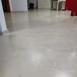 Jasnoszara posadzka z mikrocementu z widocznymi smugami i refleksami światła, w tle biurowe wyposażenie.