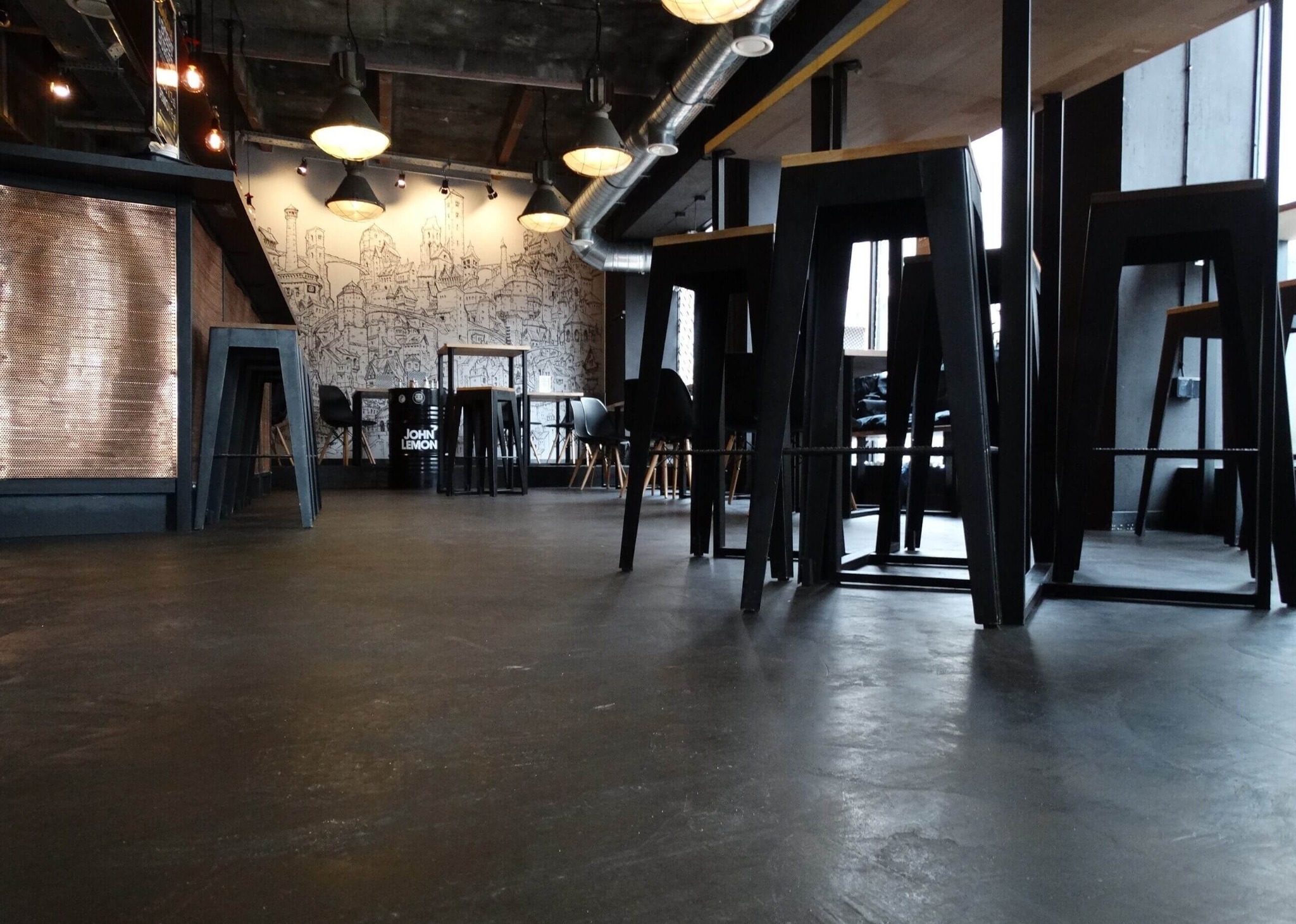 Wnętrze kawiarni z ciemną, gładką posadzką, wysokimi czarnymi stołkami barowymi i muralem na ścianie w tle.