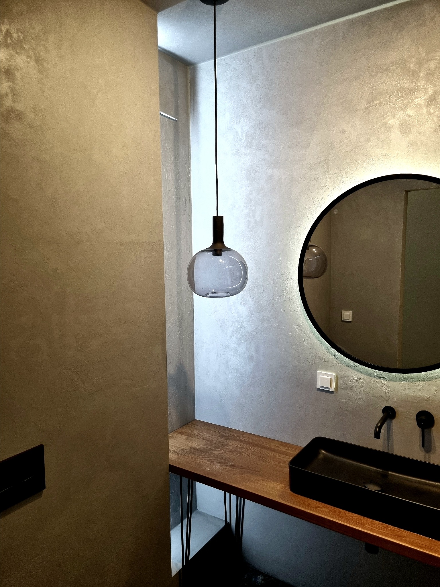 Szara ściana z efektem dekoracyjnym, okrągłe lustro z podświetleniem LED, czarna umywalka na drewnianym blacie z metalowymi nogami i wisząca szklana lampa.