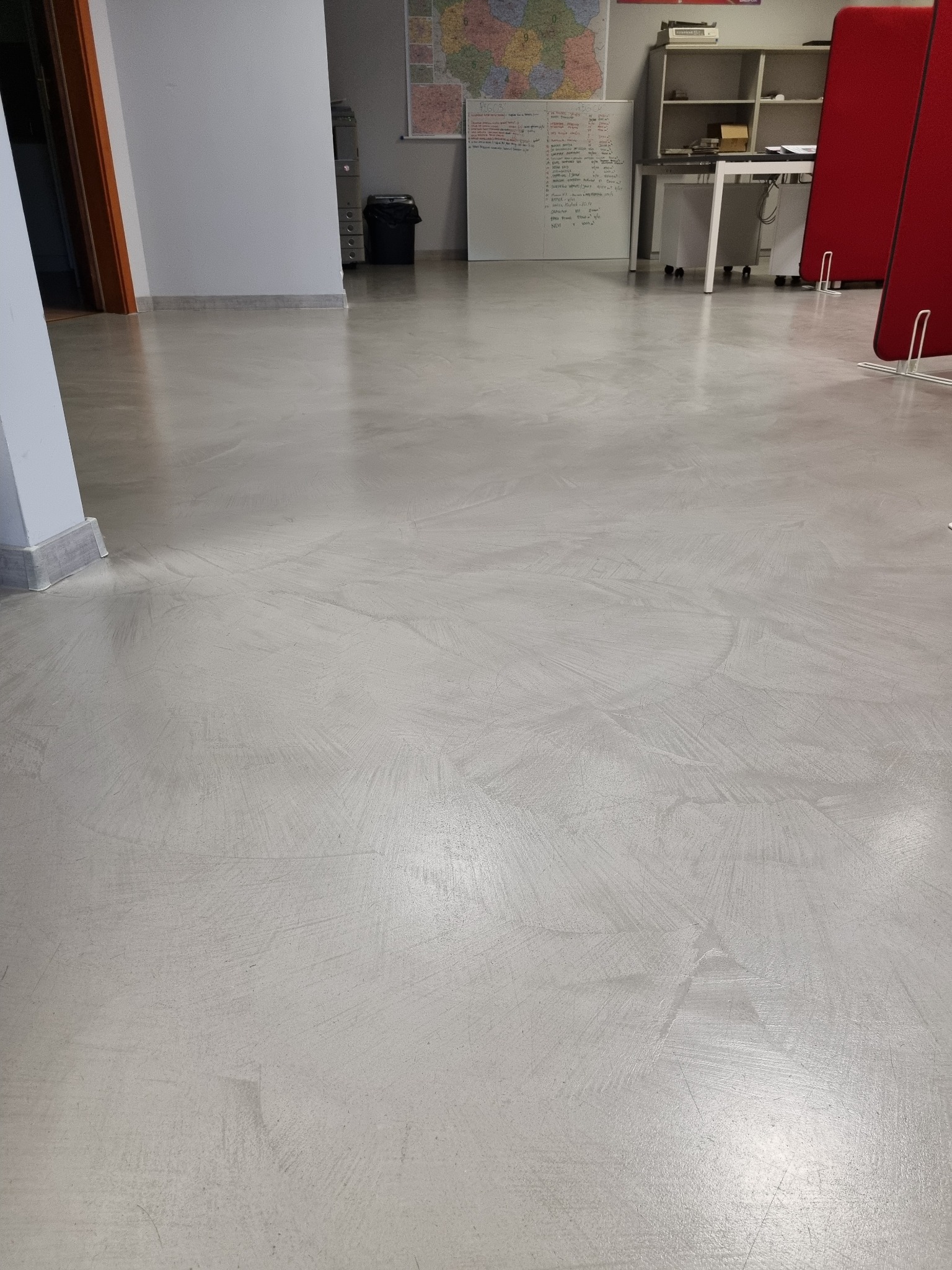 Jasnoszara posadzka z mikrocementu z widocznymi smugami i refleksami światła, w tle biurowe wyposażenie.