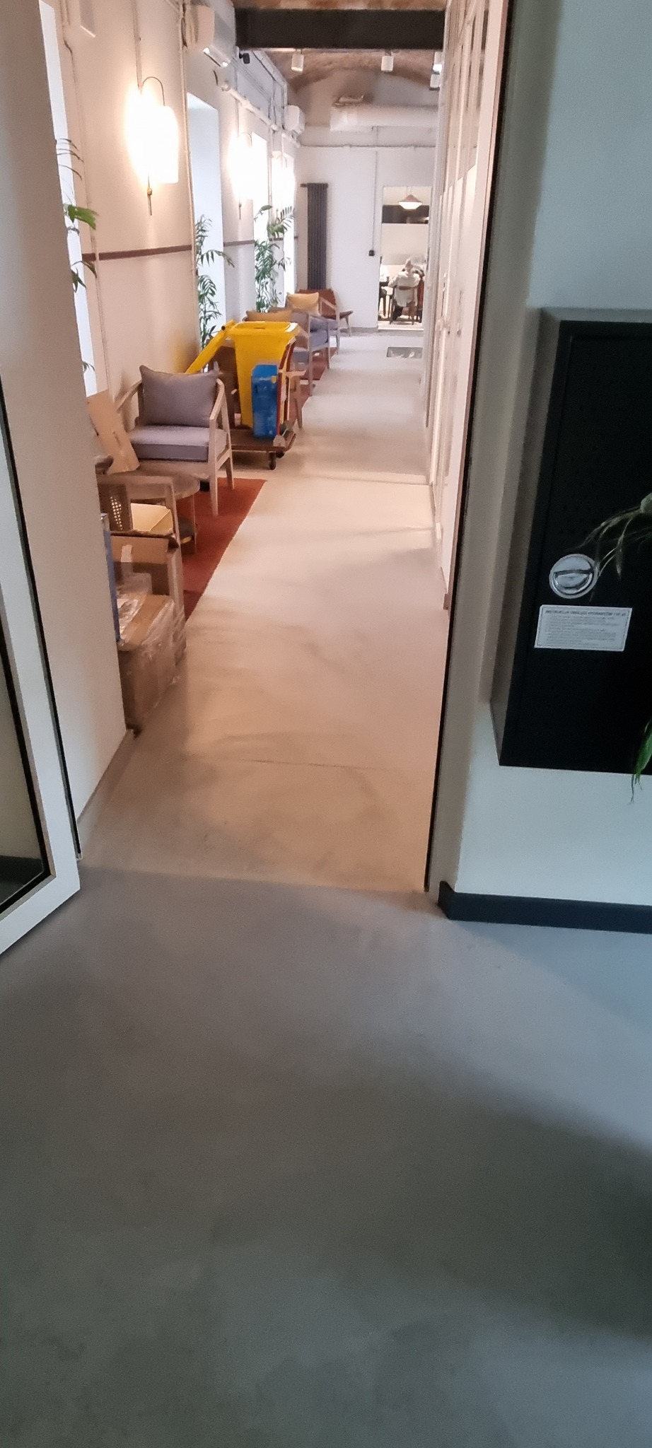 Długi korytarz z szarą posadzką, widok na wnętrze z meblami i oświetleniem, fragment szafki w prawym dolnym rogu.