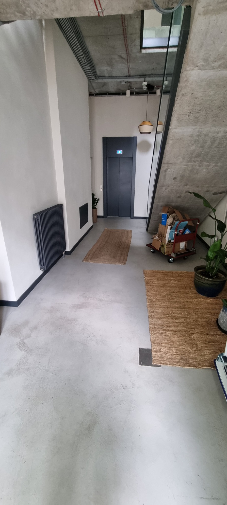 Szara posadzka w korytarzu z grzejnikiem, windą i schodami, widoczne dywaniki jutowe i wózek z kartonami.