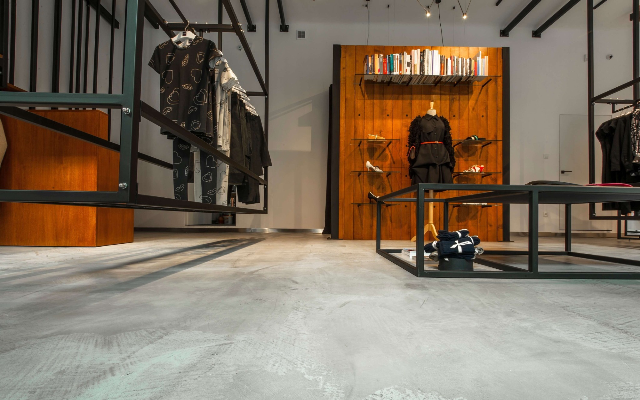 Jasnoszara posadzka z mikrocementu w butiku odzieżowym, z widoczną fakturą i delikatnymi smugami, w tle drewniana ściana z półkami na książki i ubrania na manekinie, metalowe regały z odzieżą.