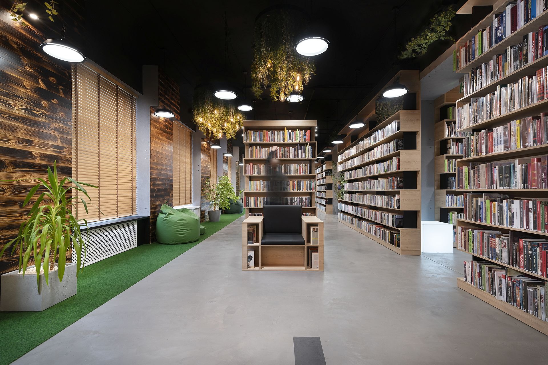 Wnętrze biblioteki z betonową posadzką, regałami z książkami, drewnianymi panelami ściennymi i zielonymi akcentami roślinnymi oraz fotel z półkami na książki.