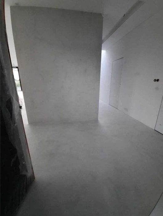 Surowe wnętrze w trakcie prac wykończeniowych: szara, gładka posadzka i ściany, częściowo zakryte folią zabezpieczającą.