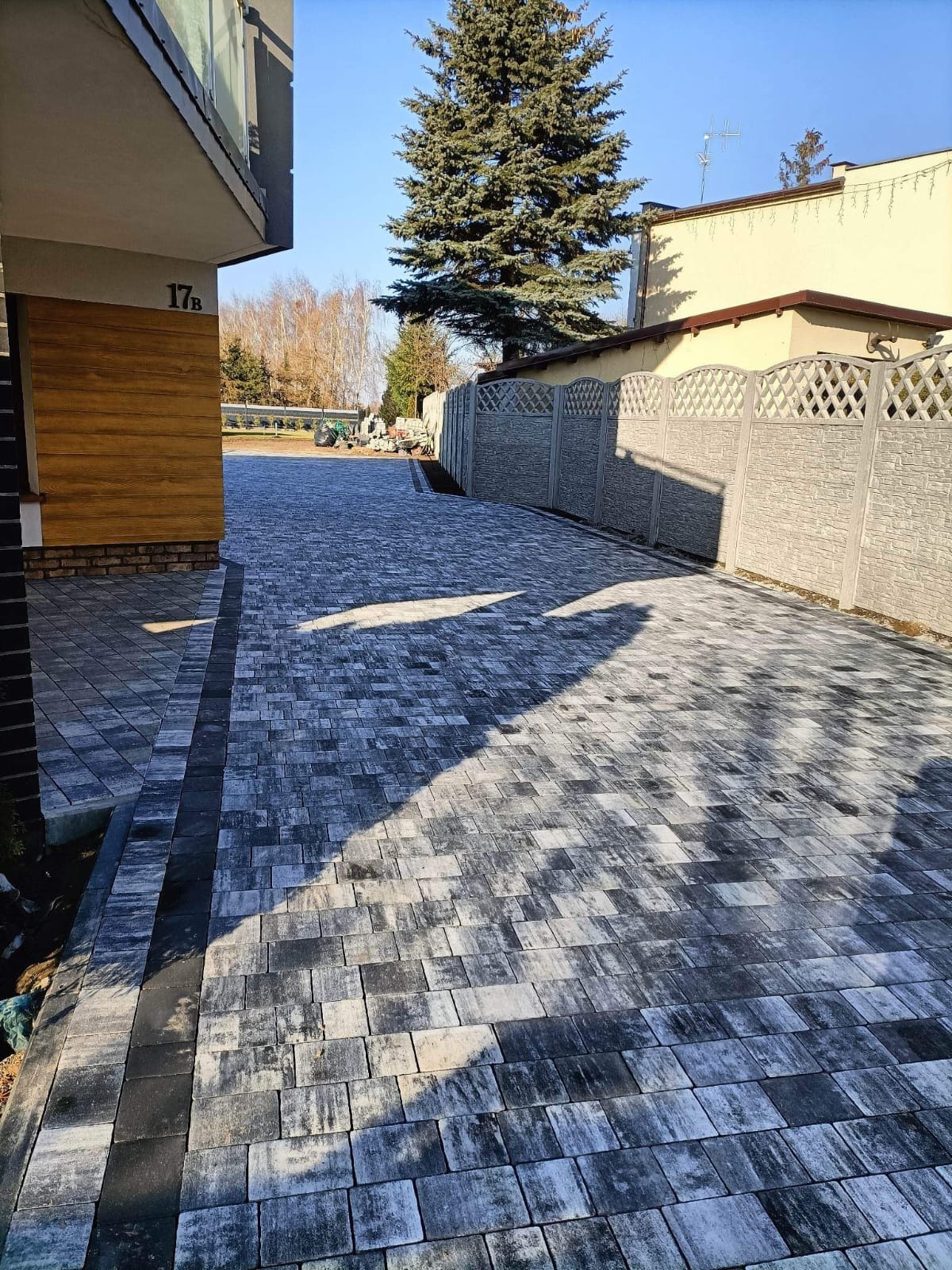 Nowo ułożona, szara kostka brukowa prowadząca wzdłuż betonowego ogrodzenia i budynku z numerem 17B w słoneczny dzień.
