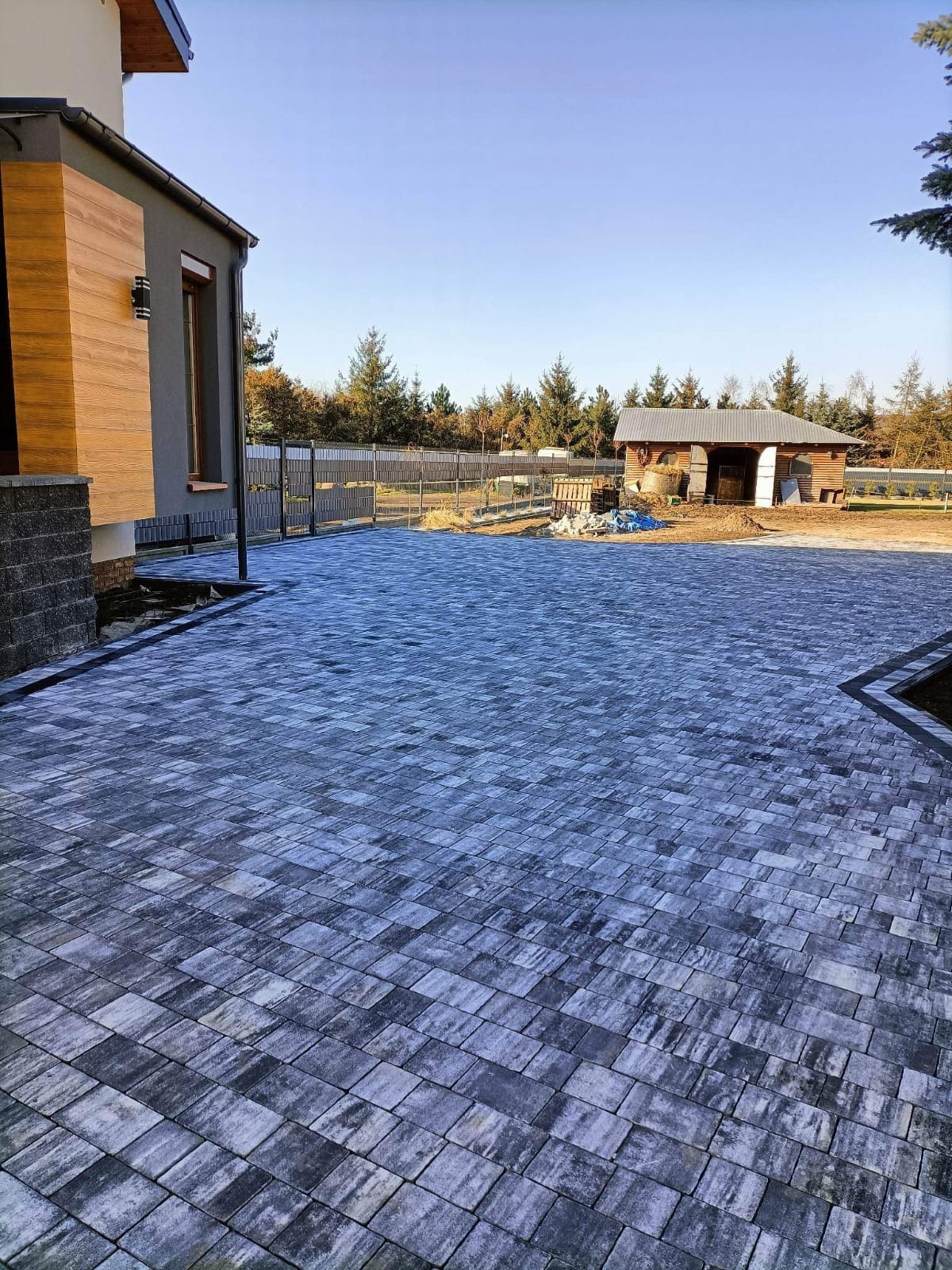 Ułożona szara kostka brukowa na podjeździe przed domem z elementami drewnianej elewacji i stajnią w tle, widok z poziomu gruntu.