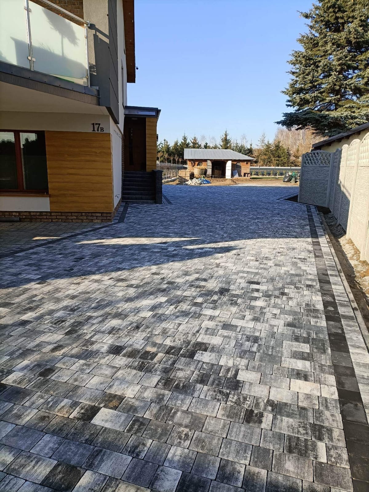 Ułożona szara kostka brukowa na podjeździe w Miłosławiu, widok na dom z numerem 17b i budynek gospodarczy w tle.