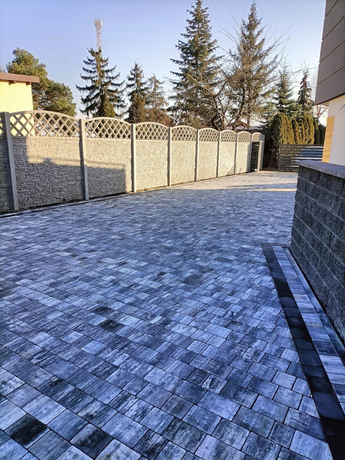 Wykończony podjazd z szarej kostki brukowej ułożonej w regularny wzór, z betonowym ogrodzeniem z dekoracyjnym wzorem i widocznymi drzewami w tle.