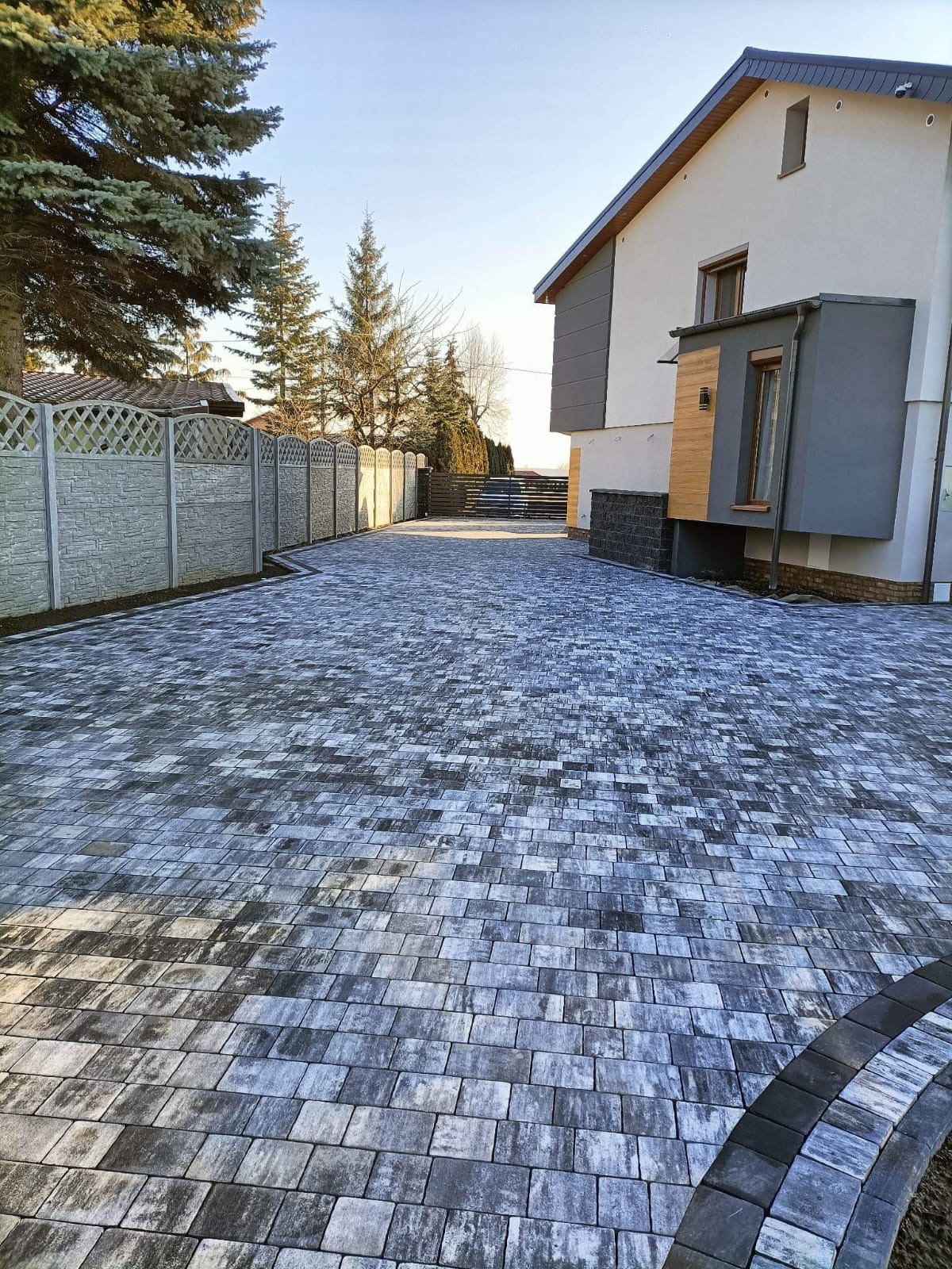 Ułożona kostka brukowa w odcieniach szarości tworząca podjazd do nowoczesnego domu z szarym ogrodzeniem panelowym. W tle widoczne drzewa iglaste i błękitne niebo.
