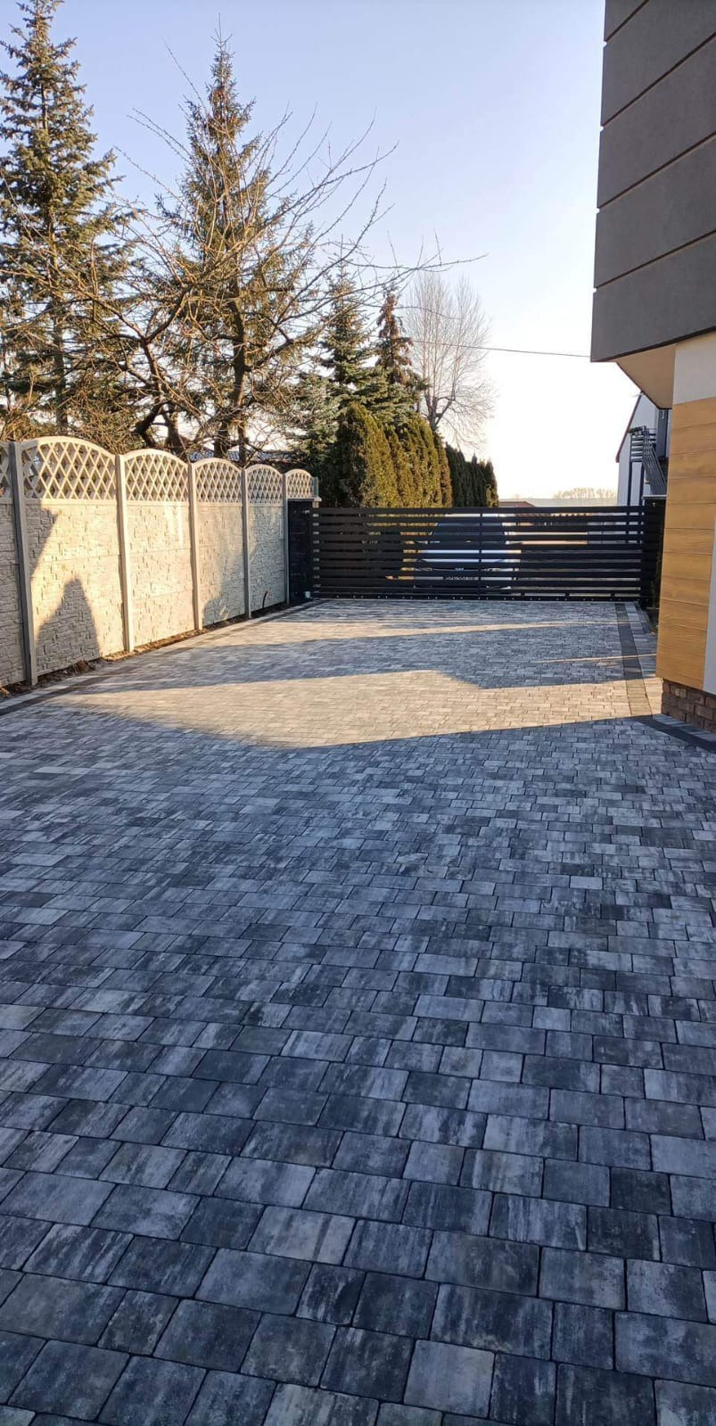 Ułożona, szara kostka brukowa na podjeździe, z widocznym ogrodzeniem betonowym z dekoracyjnym wzorem i metalową bramą przesuwną w tle, oświetlona porannym słońcem.