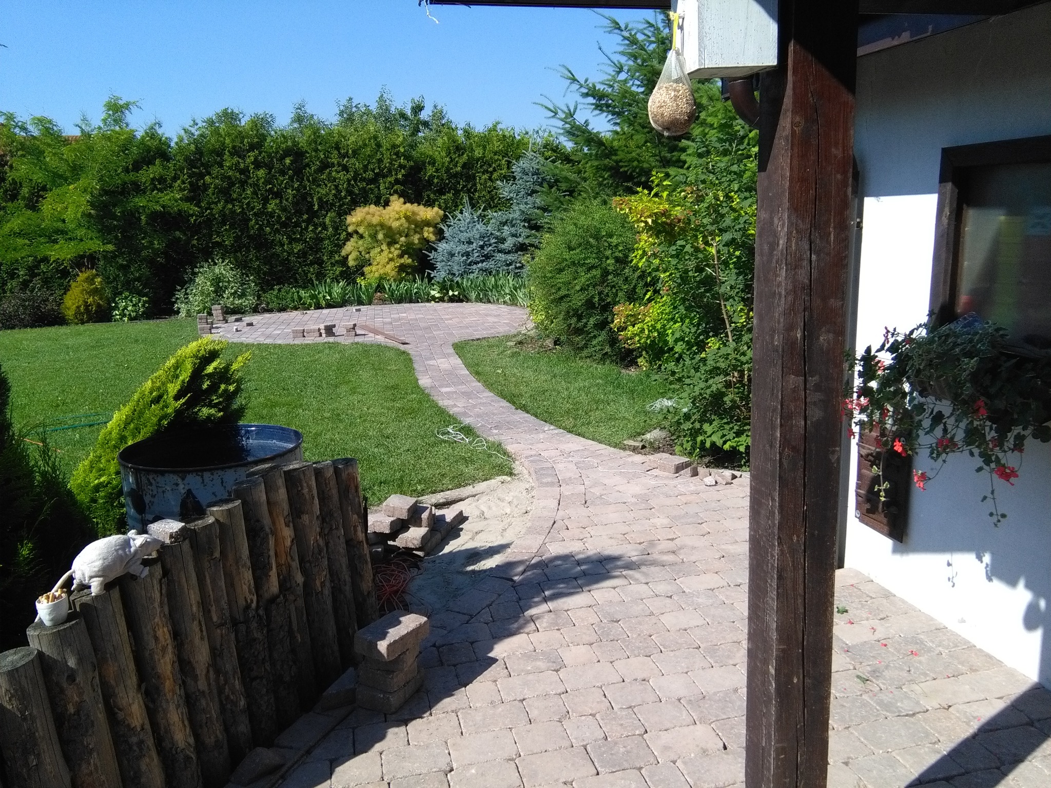 Kręta ścieżka z jasnej kostki brukowej prowadząca przez zielony trawnik w ogrodzie, widok z zadaszonej werandy z drewnianym słupem, w tle różnorodne krzewy i drzewa.