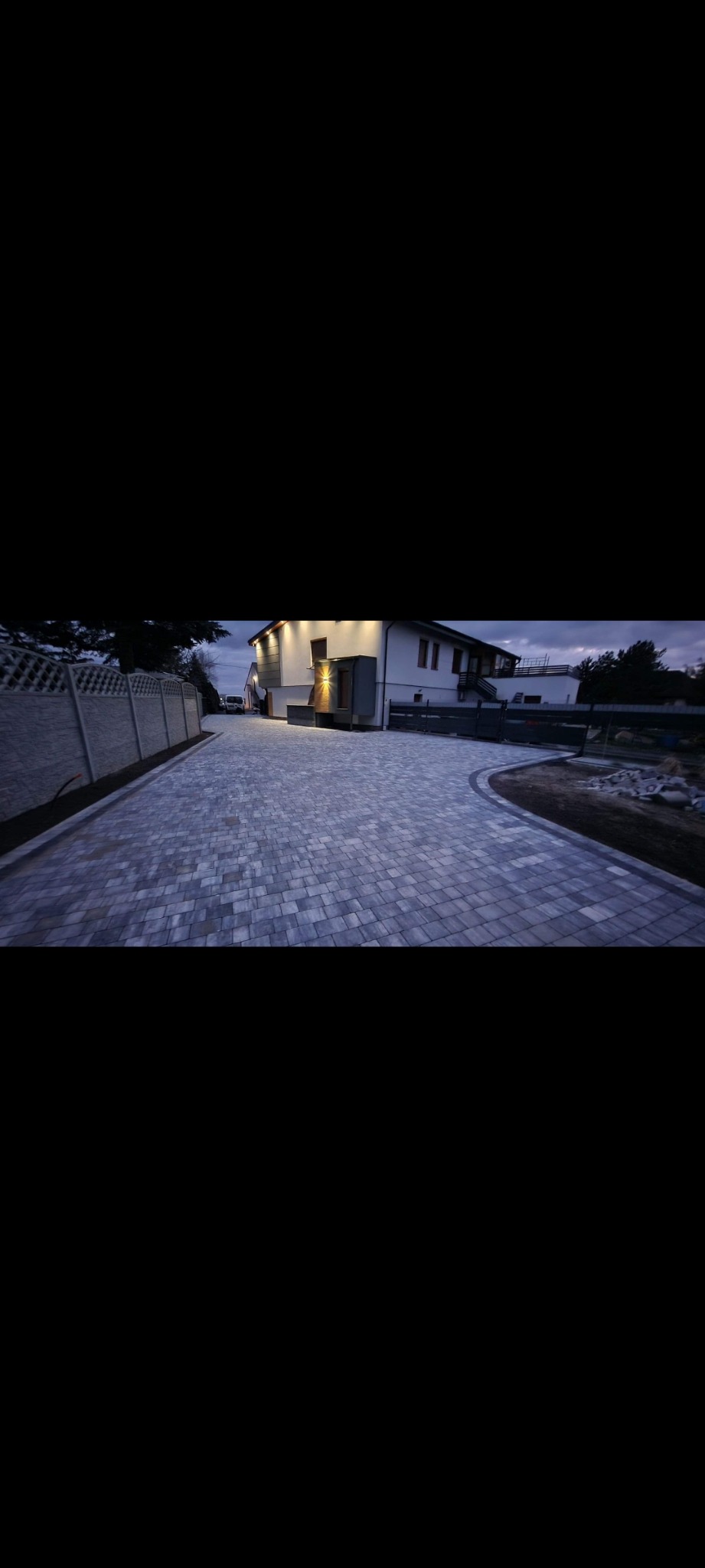 Nowoczesny podjazd z szarej kostki brukowej prowadzący do oświetlonego domu z białymi ścianami i szarymi elementami wykończeniowymi, widoczny betonowy płot po lewej stronie.