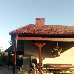 Brązowy dach z rynnami na zadaszonym tarasie, widok na komin i fragment ogrodu w słoneczny dzień.