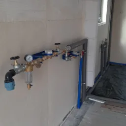 Nowo zainstalowany, niebieski panel z zaworami, filtrem i manometrem, przymocowany do surowej ściany z betonu komórkowego, widoczne rury grzewcze w izolacji.