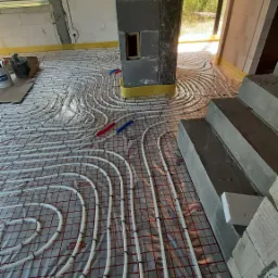 Białe rury ogrzewania podłogowego ułożone na folii refleksyjnej z czerwoną siatką, przymocowane czarnymi klipsami w surowym wnętrzu budynku w trakcie budowy, widoczne betonowe schody i kolumna.