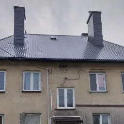 Szary, blaszany dach z dwoma kominami na dwupiętrowym budynku z kremową elewacją, widoczne okna z białymi ramami i numer budynku 18.