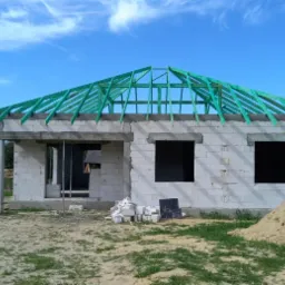 Budowa domu jednorodzinnego z widoczną, zieloną konstrukcją więźby dachowej na tle błękitnego nieba.