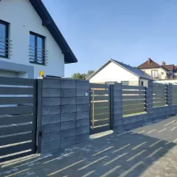 Nowoczesna, grafitowa brama przesuwna z poziomymi panelami i murkiem z szarej kostki, na tle białego domu z ciemną dachówką i błękitnego nieba.