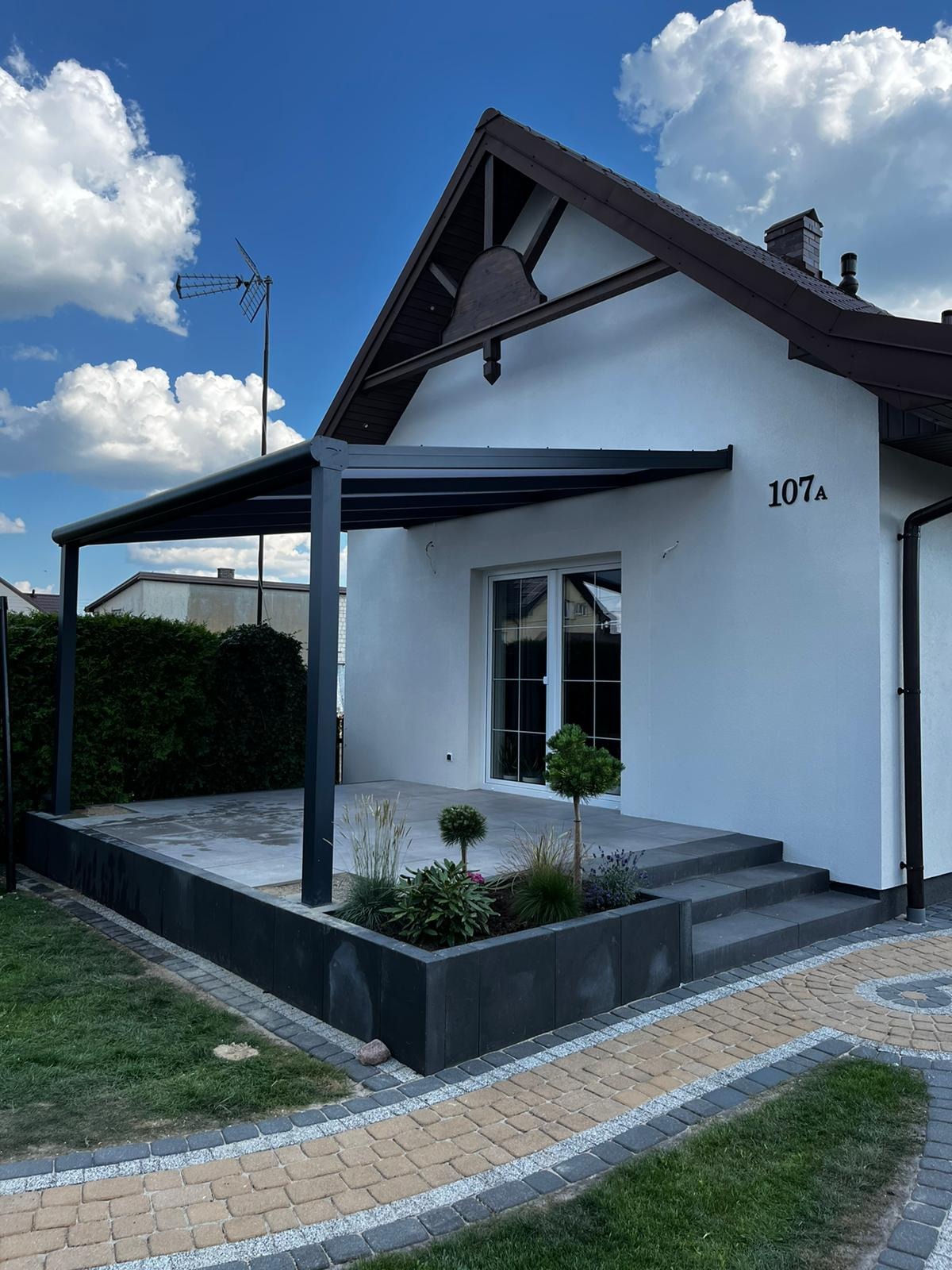 Nowoczesna, antracytowa pergola przy białym domu z tarasem i schodami, ozdobna rabata kwiatowa, kostka brukowa w dwóch kolorach, błękitne niebo z chmurami.