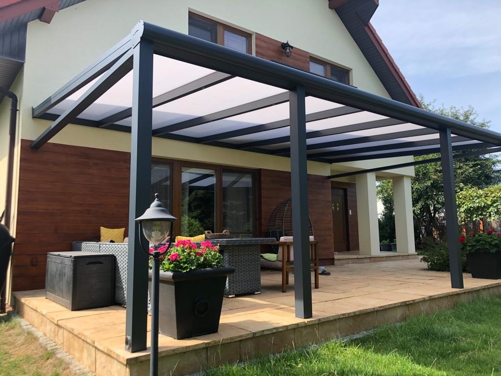 Zadaszony taras z czarną metalową konstrukcją i przezroczystym dachem, meble ogrodowe z szarej plecionki, lampa stojąca i donica z różowymi kwiatami na pierwszym planie, dom z drewnianą elewacją...