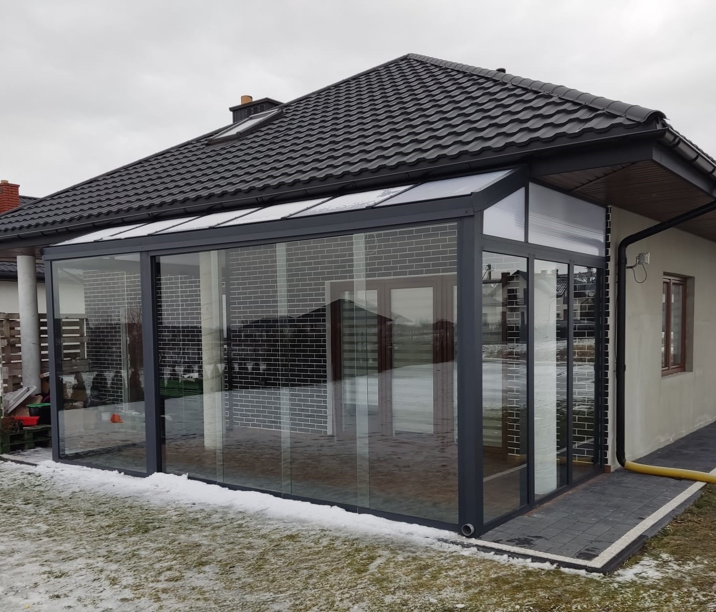 Szklana zabudowa tarasu z szarymi ramami, przylegająca do budynku z ciemną dachówką, widoczna z zewnątrz w zimowej scenerii z resztkami śniegu.