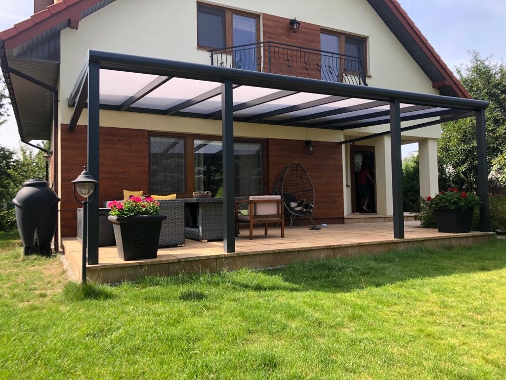 Nowoczesna pergola metalowa z przezroczystym zadaszeniem, zintegrowana z tarasem wyłożonym jasnymi płytkami, zestaw mebli ogrodowych z ciemnego rattanu, widoczny zbiornik na deszczówkę.