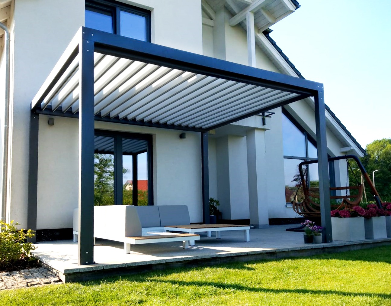 Nowoczesna metalowa pergola z regulowanymi lamelami, szara sofa i stolik kawowy na betonowym tarasie przy białym domu z dużymi oknami, huśtawka ogrodowa w tle.