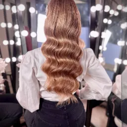 Widok z tyłu na długie, falowane, miedziano-blond włosy z ozdobą z pereł, biała bluzka z bufiastymi rękawami, czarne dżinsy, w tle lustro z oświetleniem